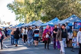 Outdoor market with blue tents and shoppers walking along a tree-lined street