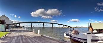 A bridge arches over water under a blue sky with fluffy clouds. Dock and building visible.