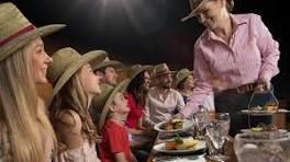 People seated at a table, served by a person holding food. All wear cowboy hats.