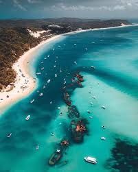 Aerial view of turquoise water with boats, along a sandy beach and shoreline with green trees under a cloudy sky.