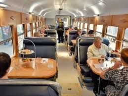 Interior of a dining car with people seated at tables, eating. Wooden paneling and windows visible.