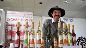 Man with mustache stands behind a display of fruit wines, including bottles of 