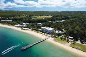 Beach scene with pier, turquoise water, white sand, buildings, and green forest in the background.