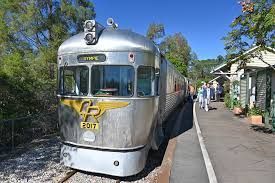 Silver train at a station with people nearby, under a bright blue sky.