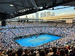 Tennis match in a packed stadium; blue court, spectators in bleachers, sunlight.