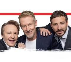 Three men smiling, posing. One in a white t-shirt, two in suits, with a red stripe above.