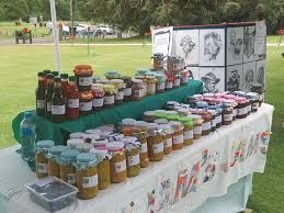 Jars of homemade preserves displayed on tables at an outdoor market, with art in the background.