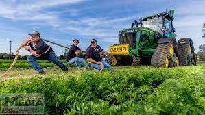 Three men struggle to pull a tractor across a field. The tractor is green and labeled 