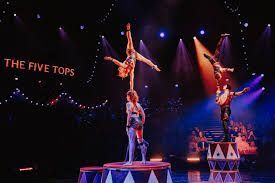 Acrobats performing a circus act. One woman balances atop two others; a second man balances on a stack of drums.