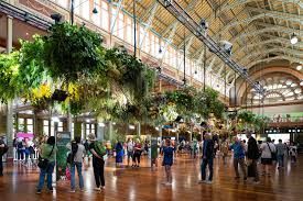 Large hall with hanging plants and people, wooden floor, arched windows.