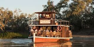 A riverboat filled with people cruises on the water, flanked by lush greenery on either side.