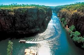 Boat navigates a narrow river canyon with steep, rocky cliffs under a bright blue sky.