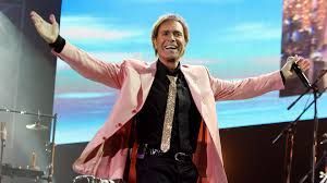 A man in a pink jacket and tie is standing on a stage with his arms outstretched.