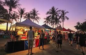 Outdoor market at dusk with vendors, customers, palm trees, and a colorful sky.