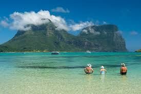 Three people wading in turquoise water at a beach with a mountain in the background.