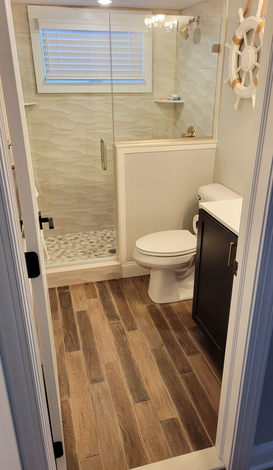Small bathroom with wood-look floor, glass shower, and white toilet. Dark vanity, beige tile, and a decorative nautical wheel are visible.