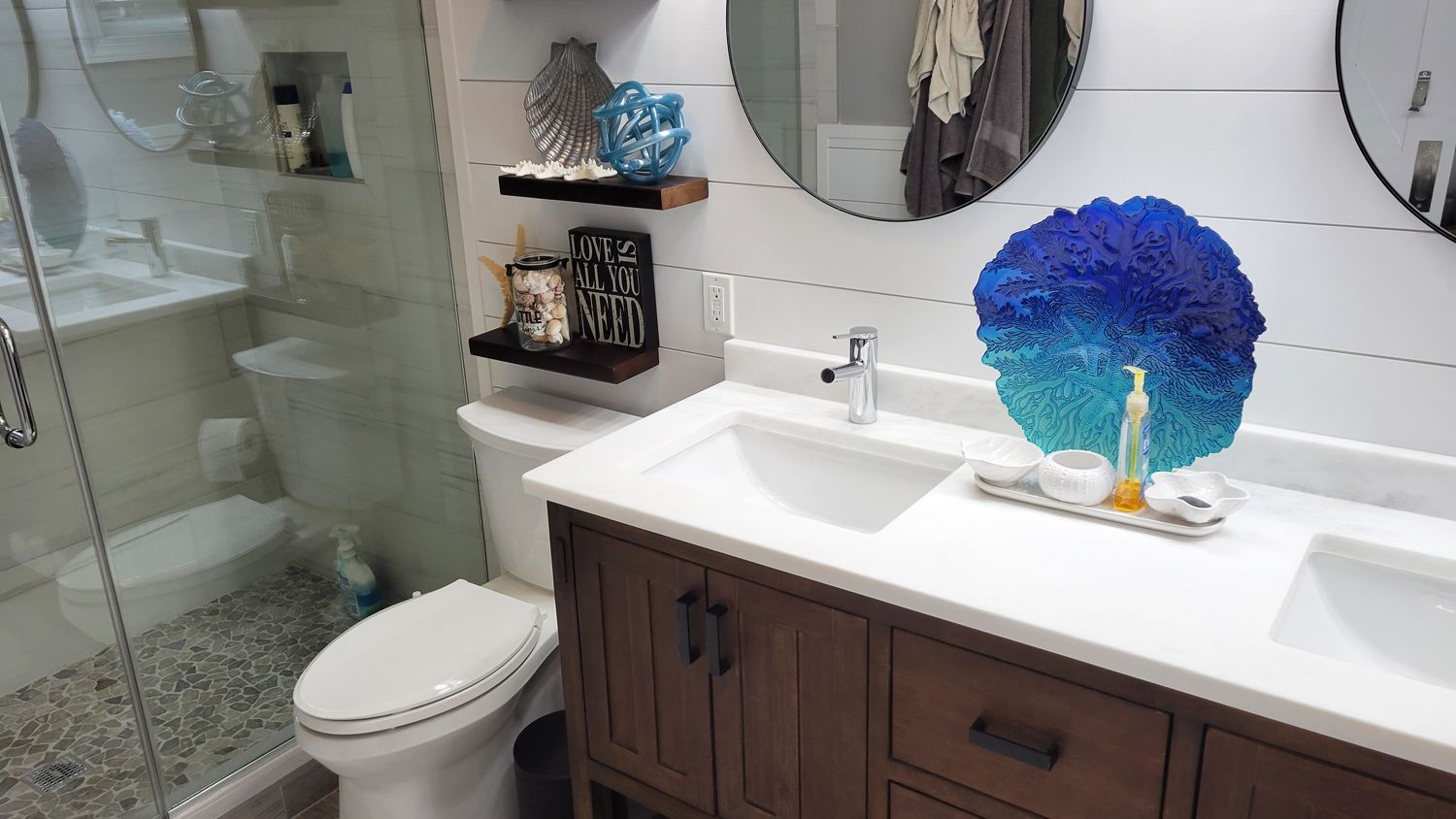 Bathroom with a dark wood vanity, white countertops, and coastal decor featuring a large blue decorative piece. A glass shower with river rock flooring is visible.