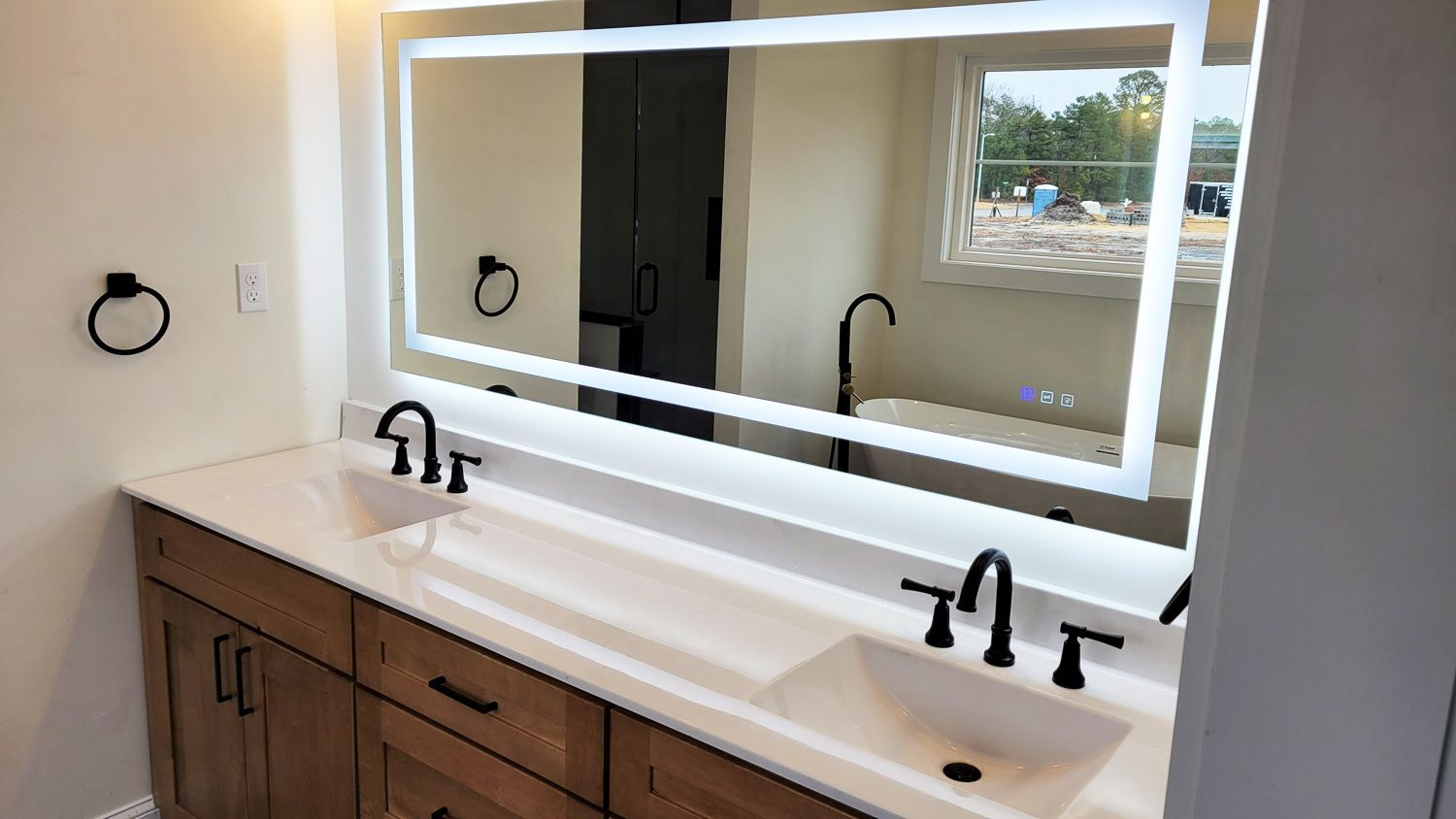 Bathroom with a double vanity, large illuminated mirror, and black fixtures. The countertop is white, and the cabinets are wood.