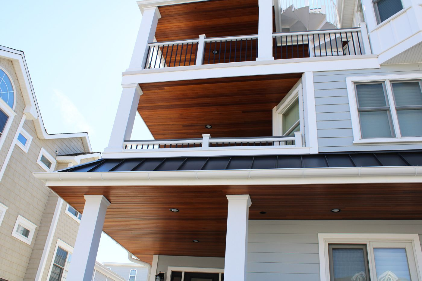 Multi-story beach house with gray siding, white trim, and brown wood ceilings on the porches; clear blue sky.