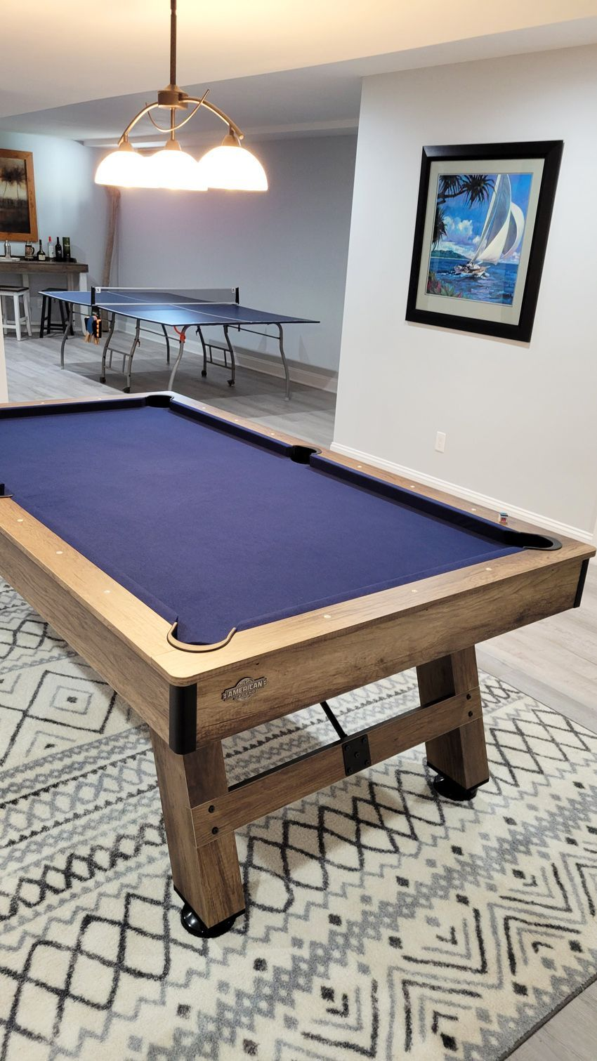 A pool table with blue felt and wooden frame in a recreation room with a ping pong table, art, and a patterned rug.