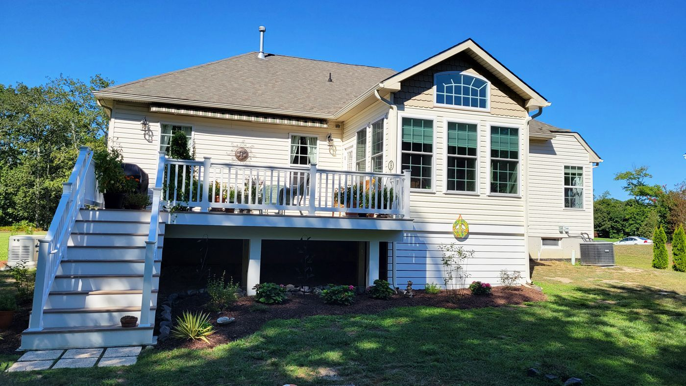 Back exterior of a light-colored house with a deck, stairs, and manicured landscaping under a blue sky.