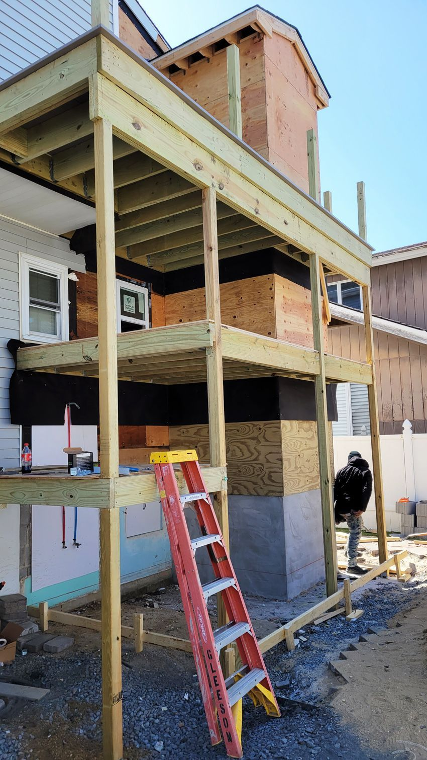 Multilevel wooden deck under construction against a building. A red ladder leans against the structure; a person stands nearby.