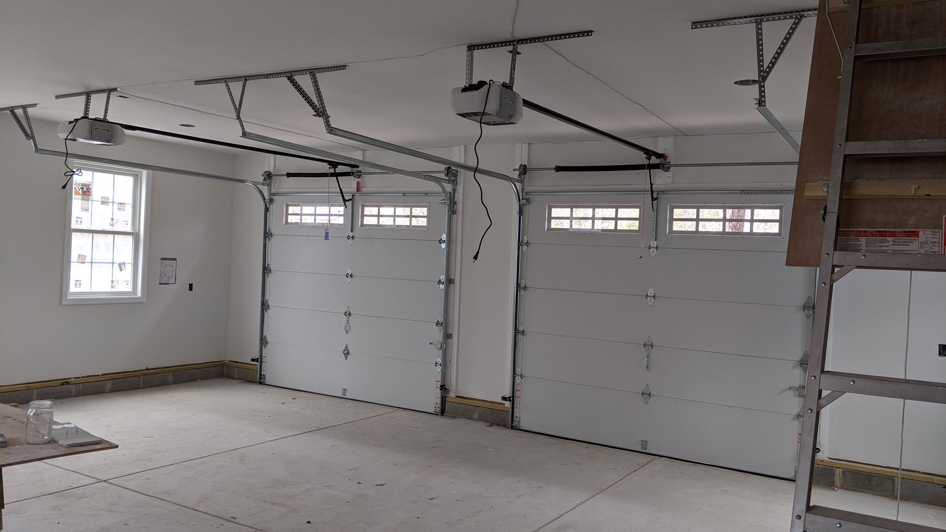 Two white garage doors with windows in a concrete garage. Overhead are two garage door openers and tracks.