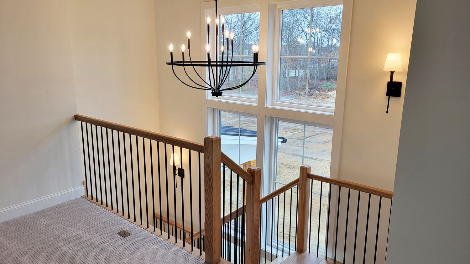 View from the top of a staircase in a home. A black chandelier hangs near large windows, with a wooden railing and carpeted floor.