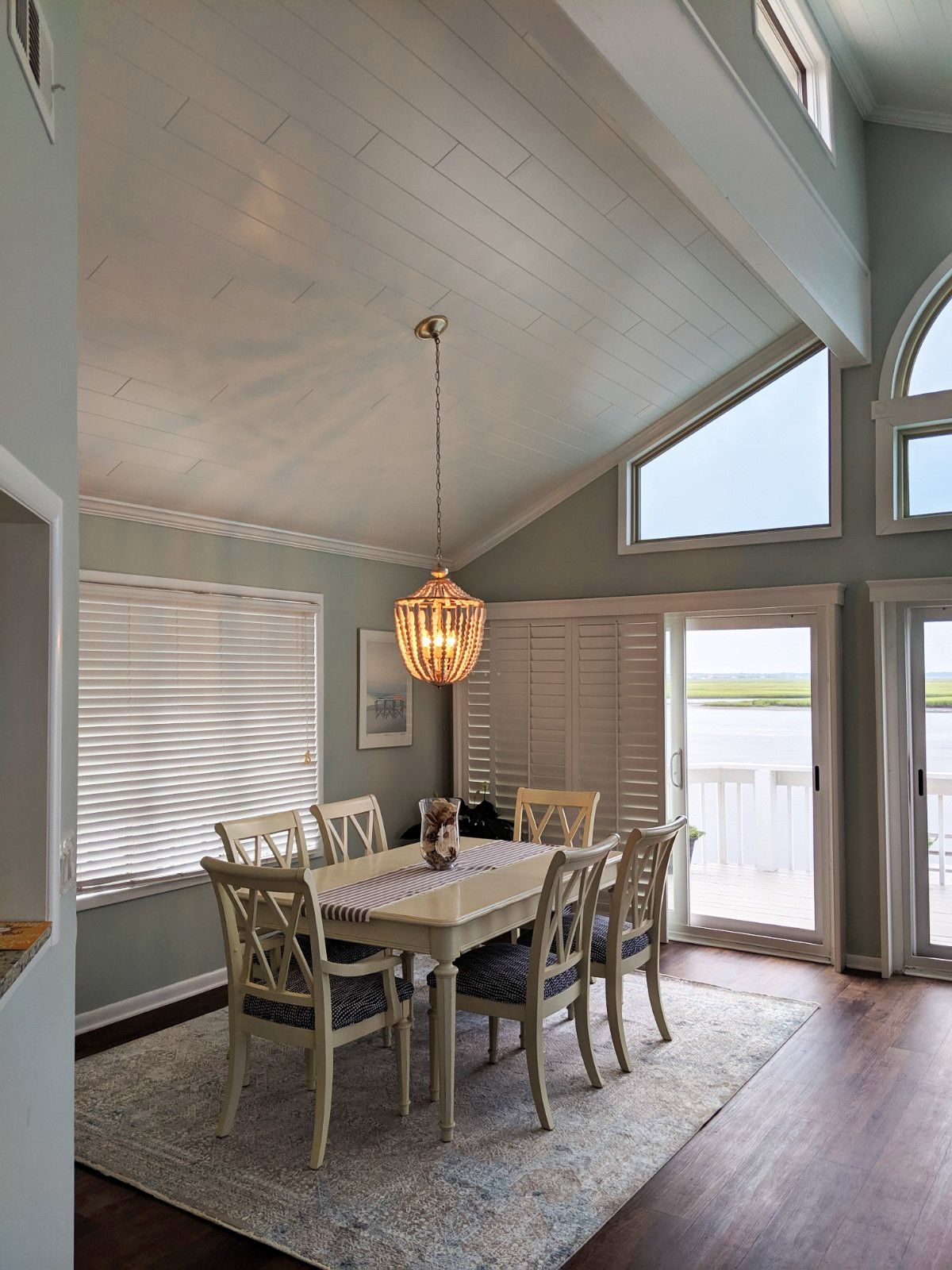 Dining room with a light-colored wooden table and six chairs on a patterned rug, under a hanging chandelier.