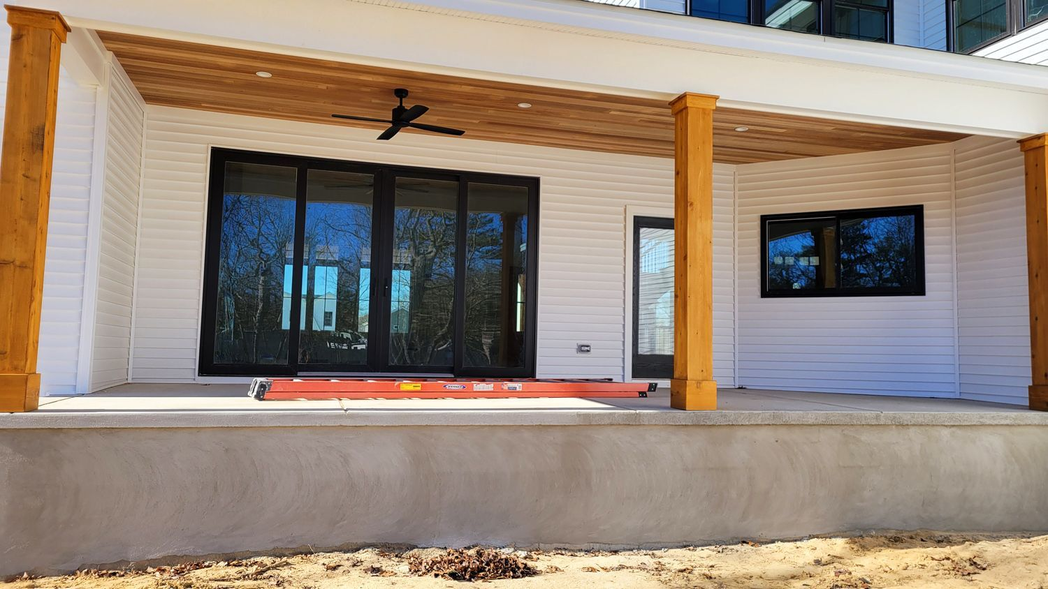 A white-sided house with a covered patio features large sliding glass doors and wooden support beams. A ceiling fan is mounted on the wood ceiling.