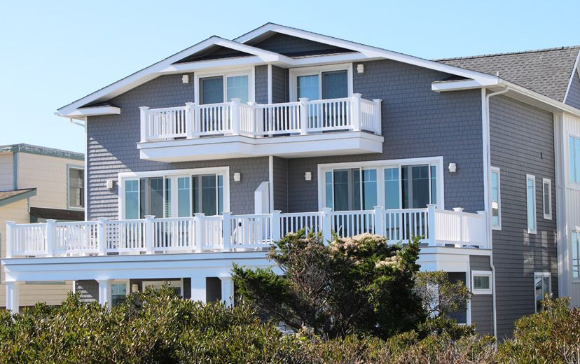Two-story gray house with white balconies and railings, overlooking some shrubs under a blue sky.