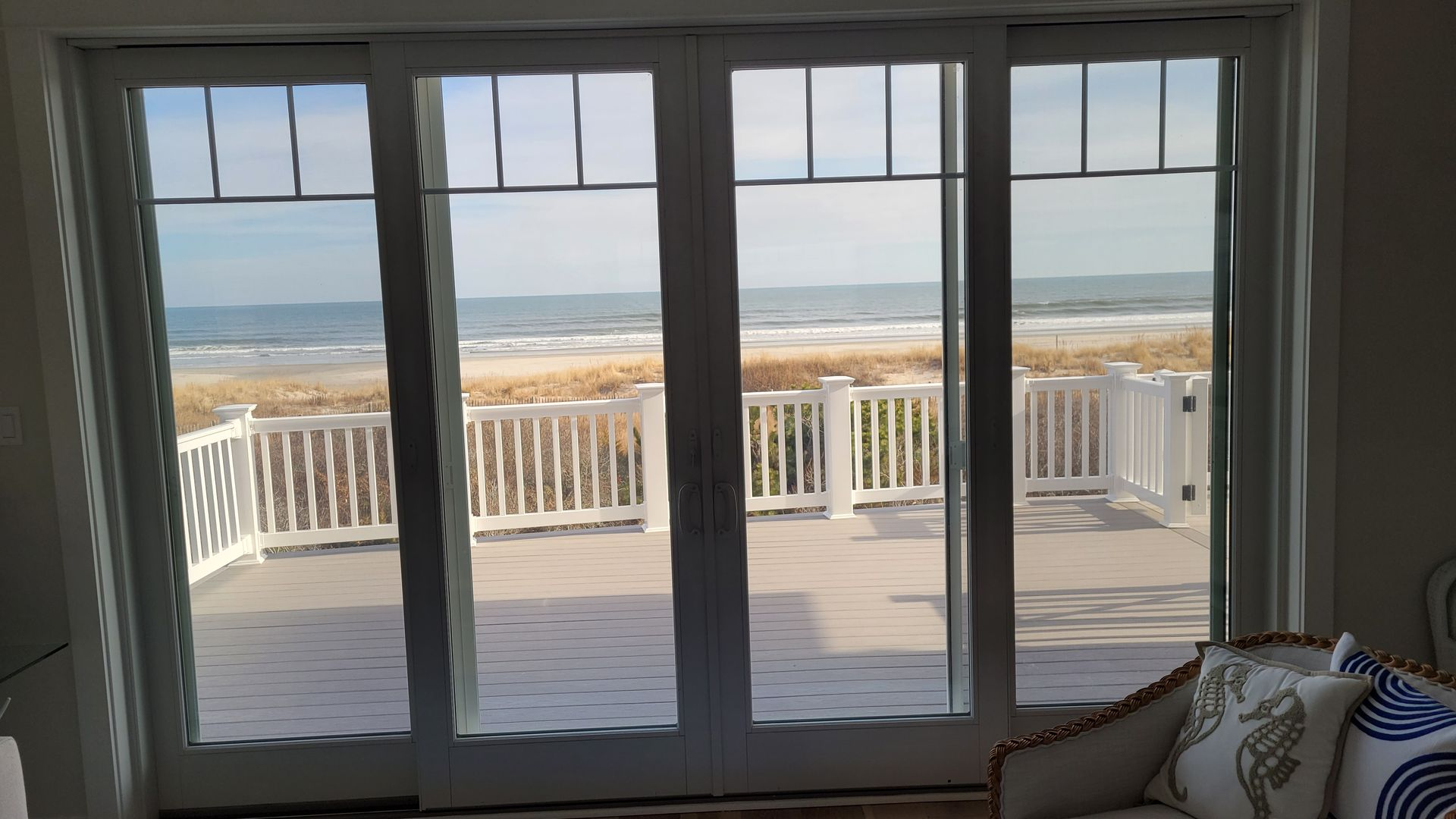 Sliding glass doors open to a beach view with a white railing, a wooden deck, and the ocean.  A sofa with pillows is in the foreground.
