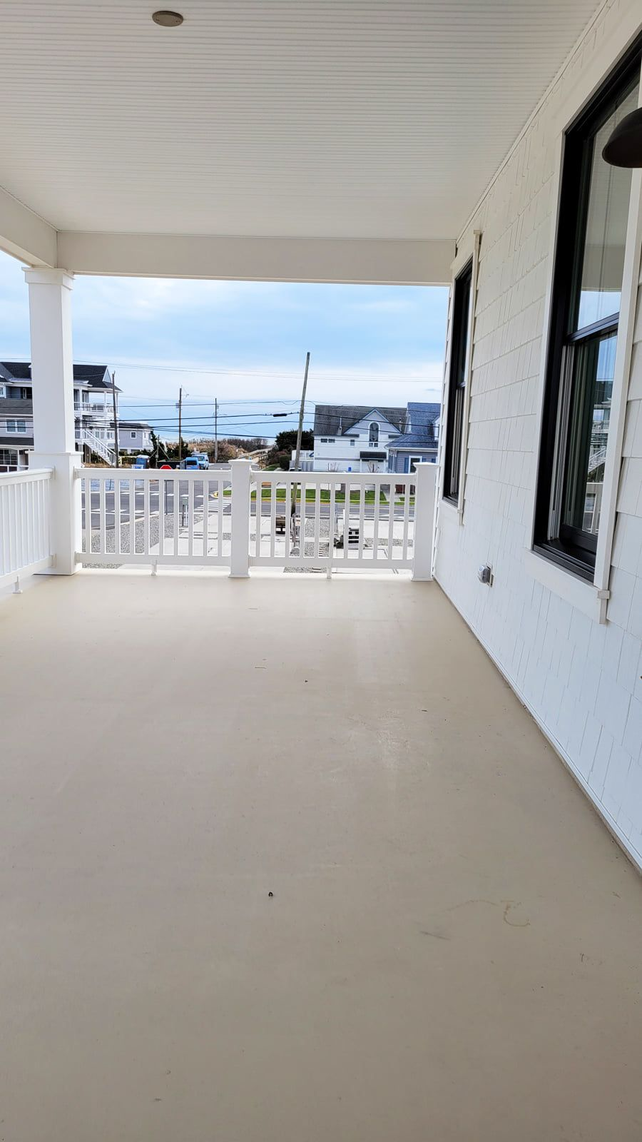 A covered porch with white railings and a beige floor overlooks a coastal scene. The sky is cloudy, with views of other buildings and the water.
