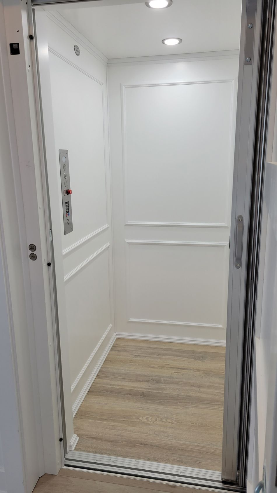 A white, interior home elevator with wood-look flooring. The open door reveals control buttons and decorative molding.