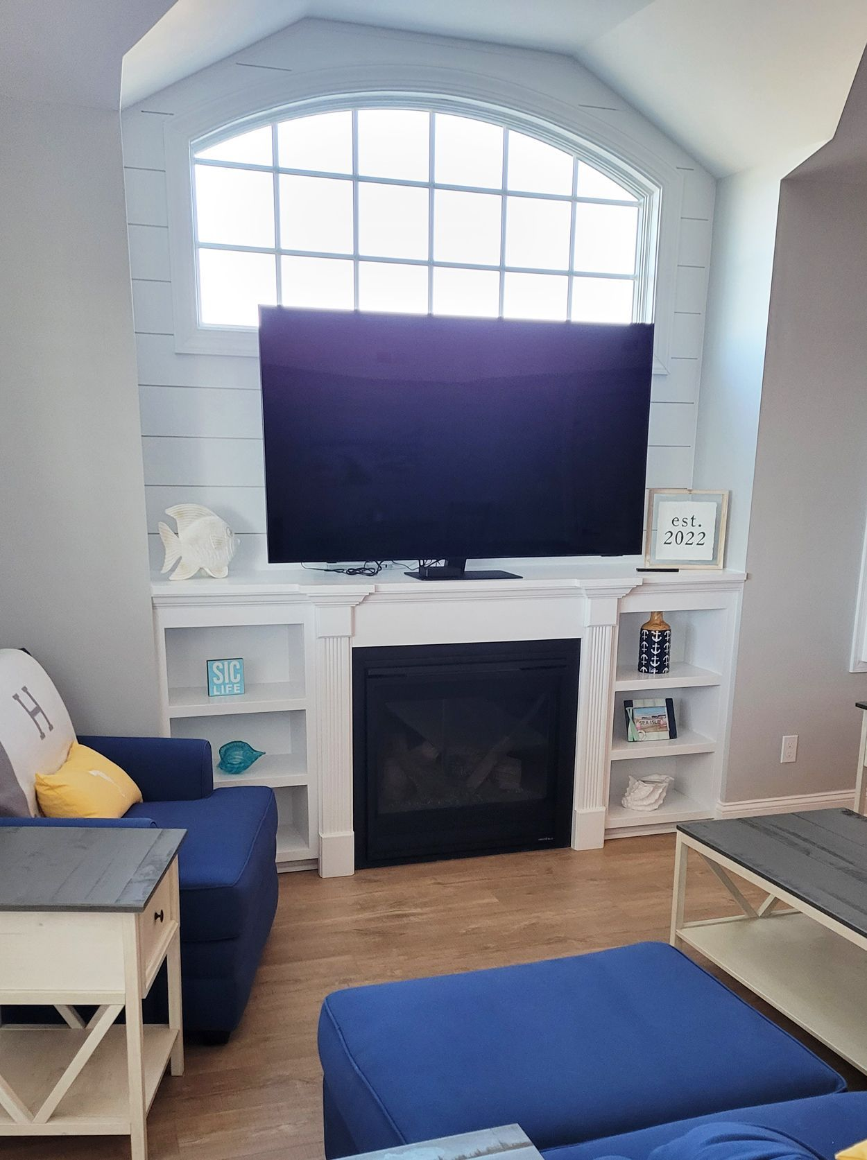 Living room with blue furniture and white fireplace with a large TV, built-in shelves, and arched window above.