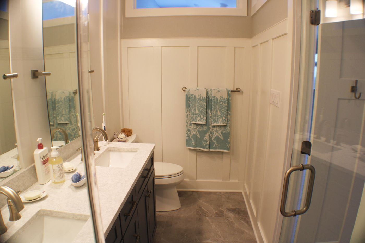 A small, modern bathroom with a vanity, toilet, and shower. Light gray walls, white wainscoting, and teal towels hang on the wall.