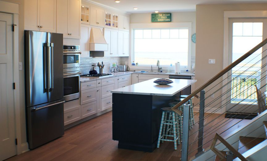 Bright kitchen with white cabinets, stainless steel appliances, and a dark blue island with a white countertop. The island has stools, and a window overlooks an exterior view.
