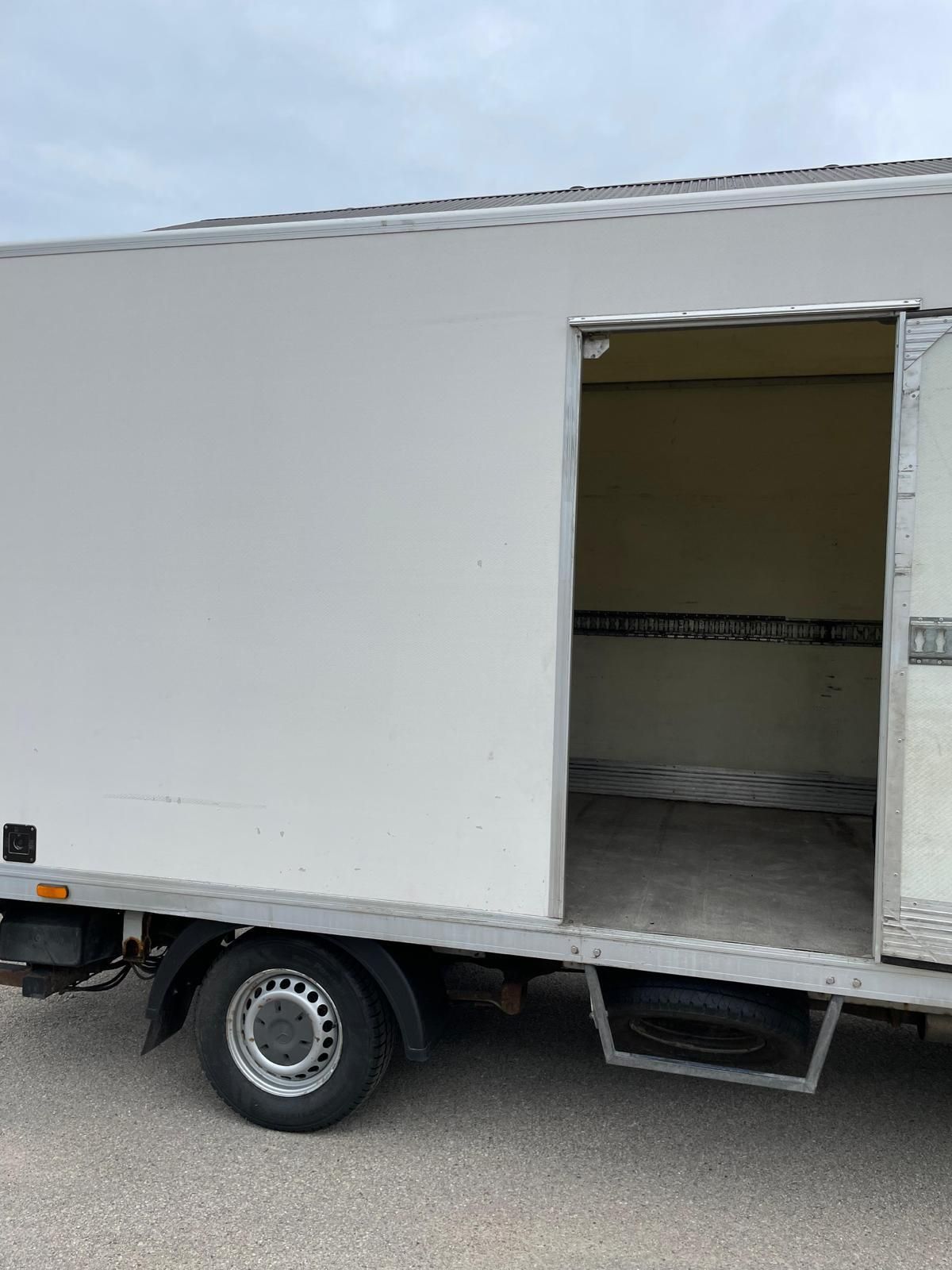 Ceļa malā stāv balta kravas automašīna ar atvērtām durvīm.