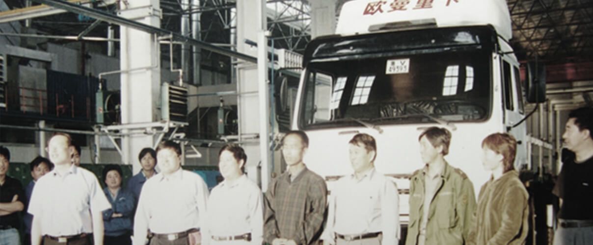 Cilvēku grupa stāv kravas automašīnas priekšā.