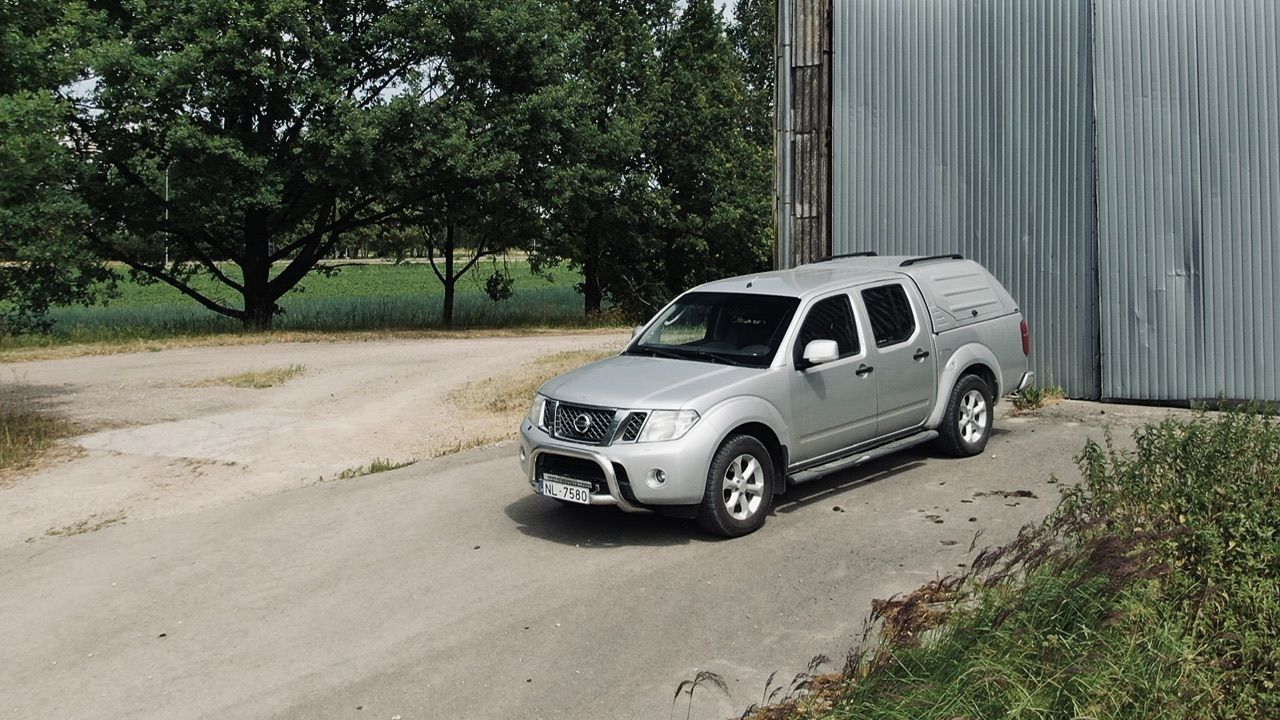 Sudraba kravas automašīna ir novietota ceļa malā pretī ēkai.