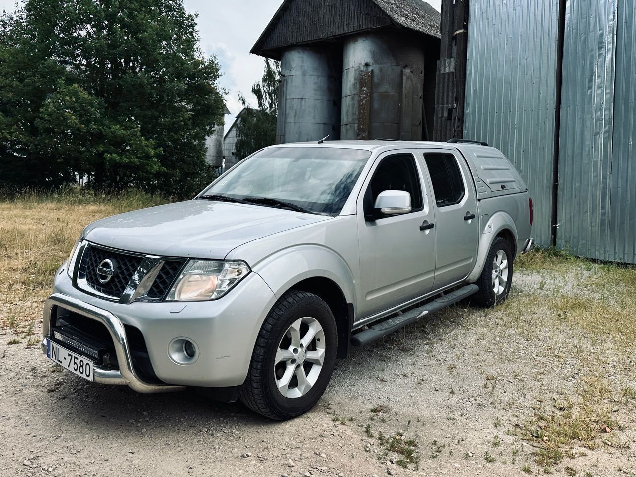 Sudraba krāsas pikaps nissan ir novietots pie kādas ēkas.