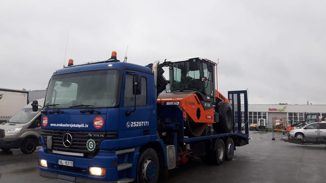 Autostāvvietā stāv zila kravas automašīna ar traktoru aizmugurē.