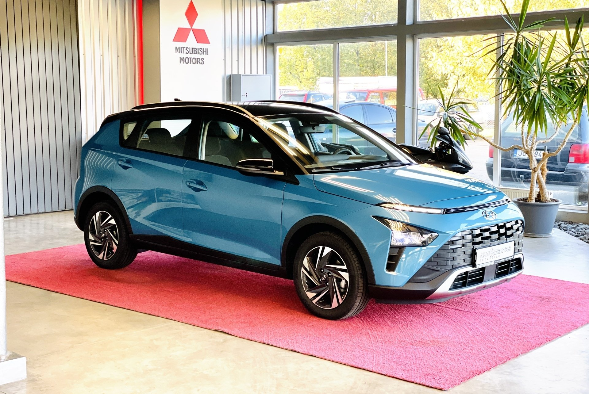 Izstāžu zālē uz rozā paklājiņa sēž zila mašīna.