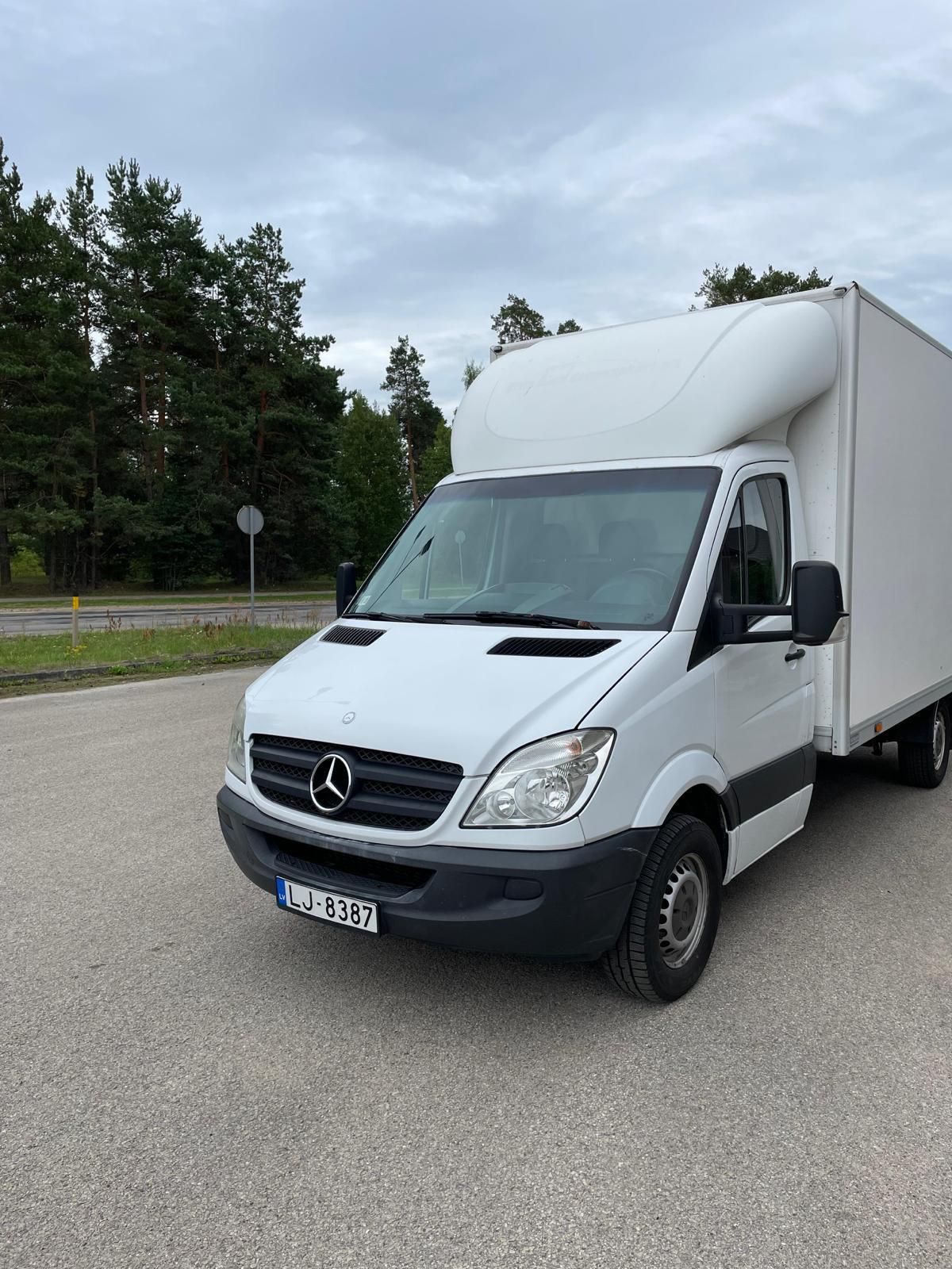 Ceļa malā stāv balts mercedes furgons.