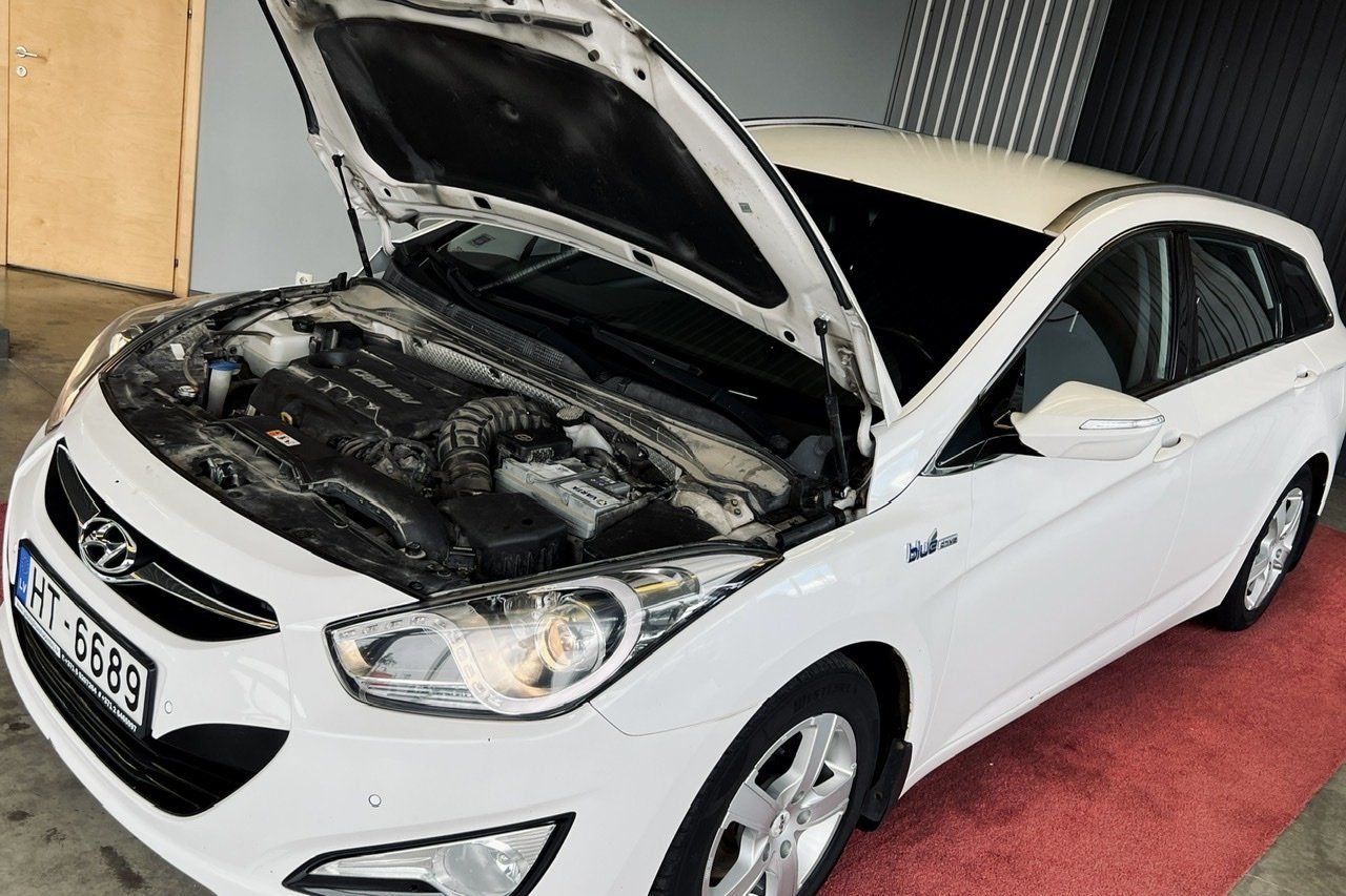 Balta automašīna ar atvērtu motora pārsegu ir novietota uz sarkanā paklāja.