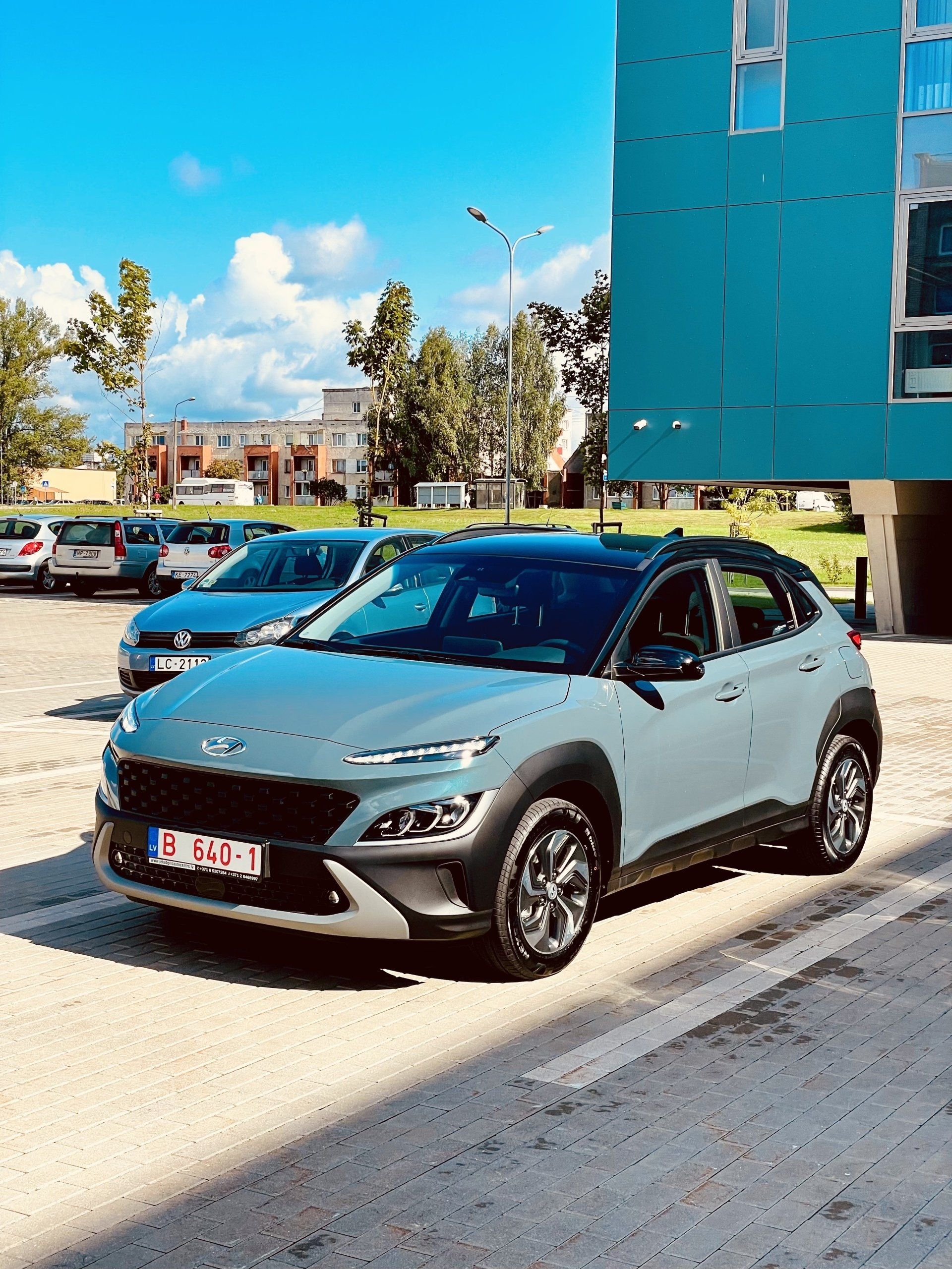 Pelēka automašīna ir novietota stāvvietā pie zilas ēkas.