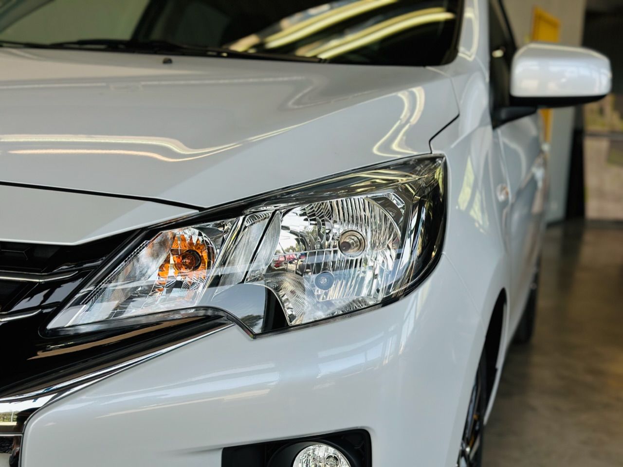 Garāžā stāv balta mašīna ar melnu motora pārsegu.