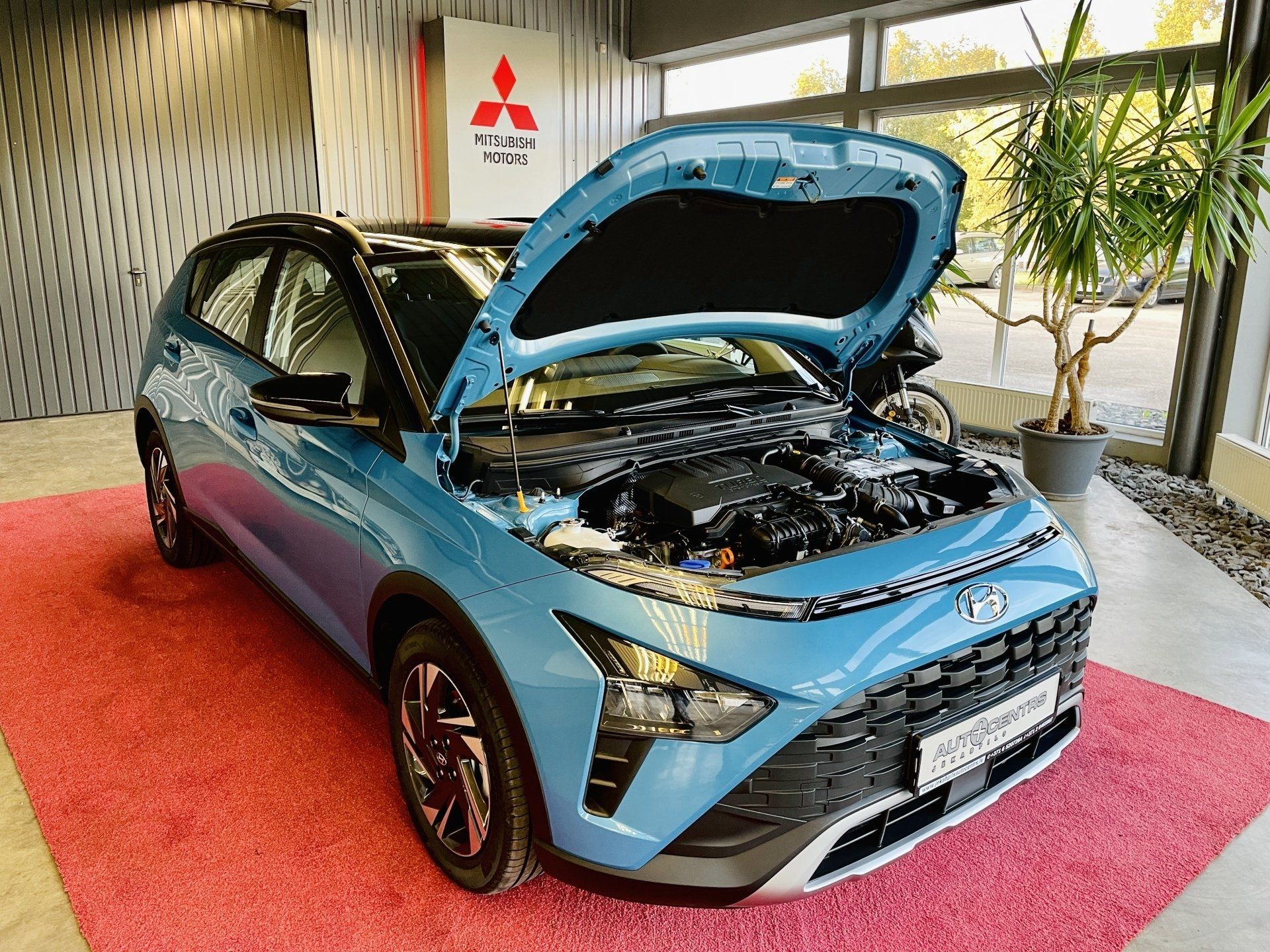 Zila mašīna ar atvērtu motora pārsegu sēž izstāžu zālē uz sarkana paklāja.