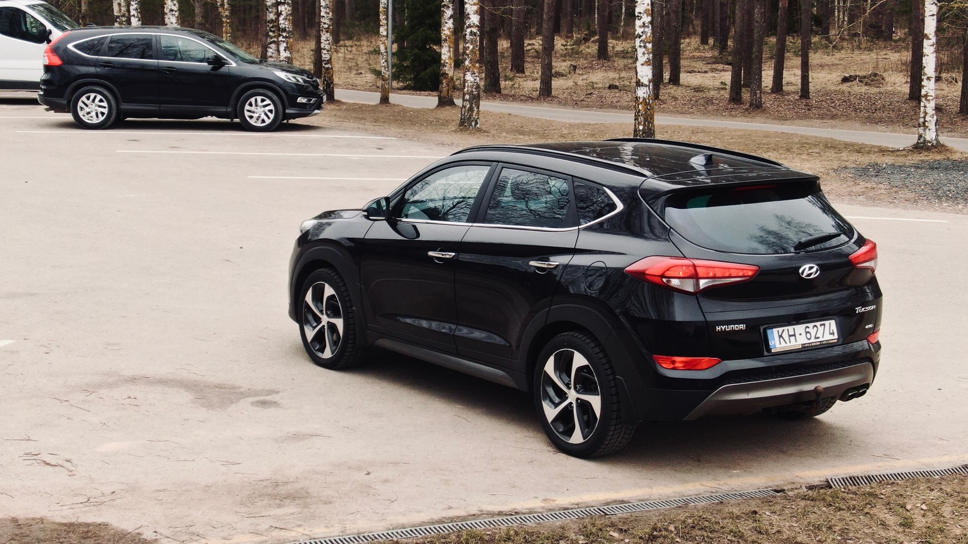 Melna mašīna stāv stāvlaukumā pie meža.