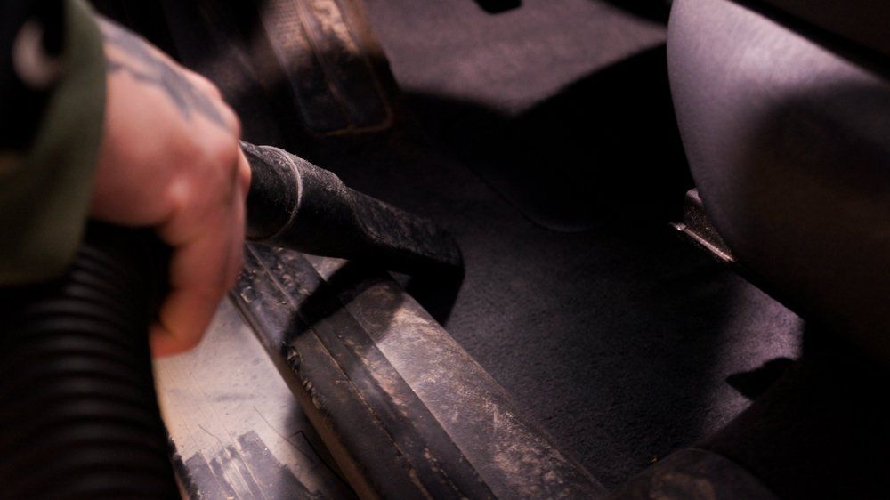 Cilvēks izmanto putekļu sūcēju, lai tīrītu automašīnas grīdu.